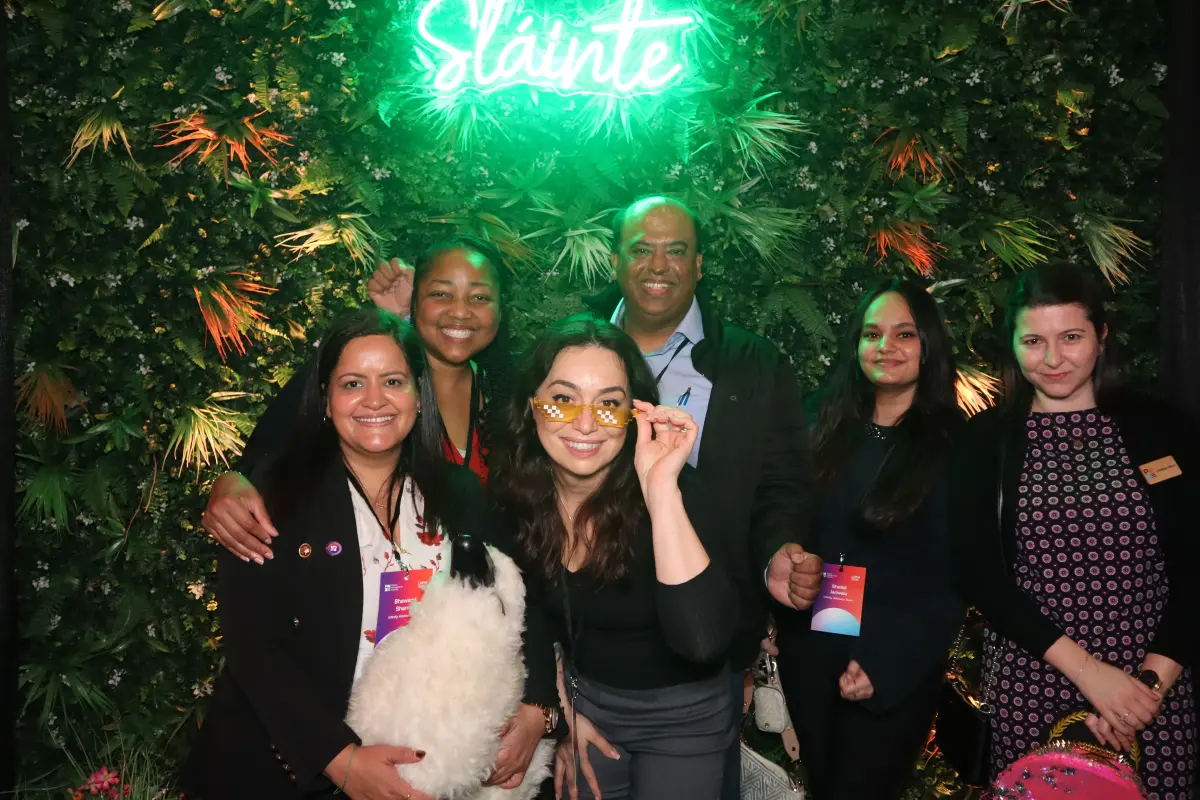 Group of friends posing at an outdoor photo booth with tropical backdrop and neon sign